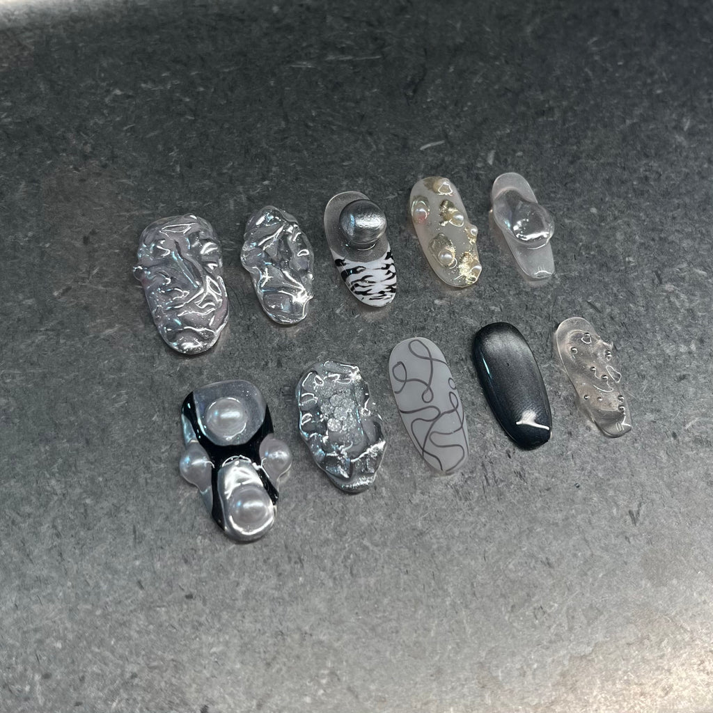 Handmade silver 3D almond press-on nails from Fjaka Nails, side-angle flat lay shot featuring raised silver texture, pearls, steel beads, clear base, zebra print, cat eye and abstract designs, salon-quality reusable nails for UK niche industrial aesthetic lovers.